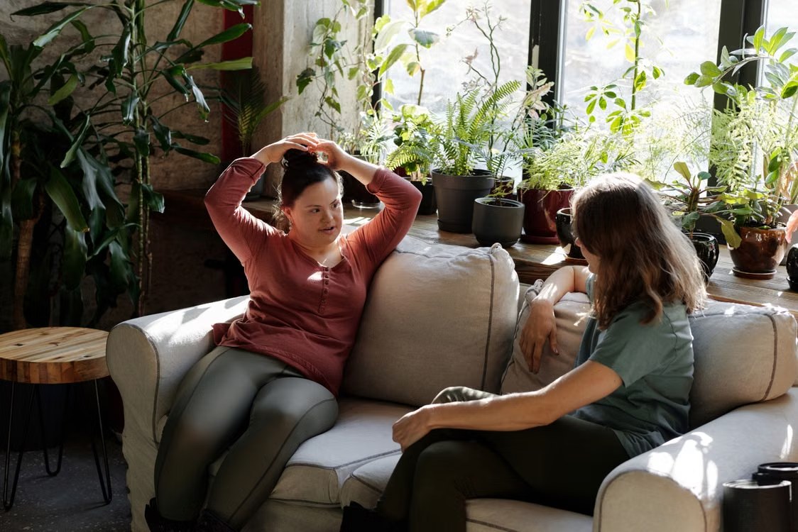 Two young women chatting on a sofa in a bright, plant-filled room