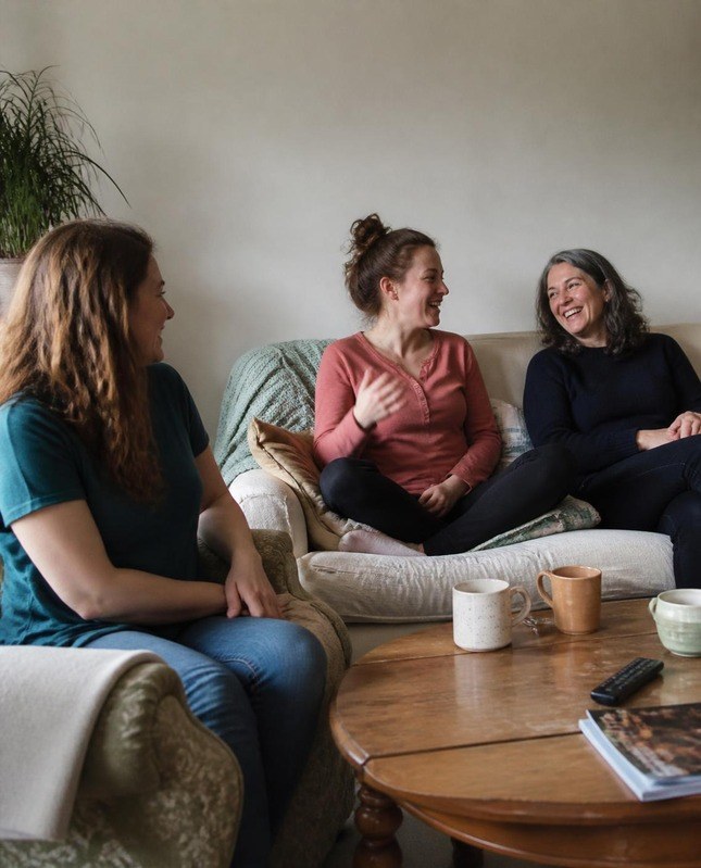 Three women laughing together on sofas around a coffee table with mugs in a living room